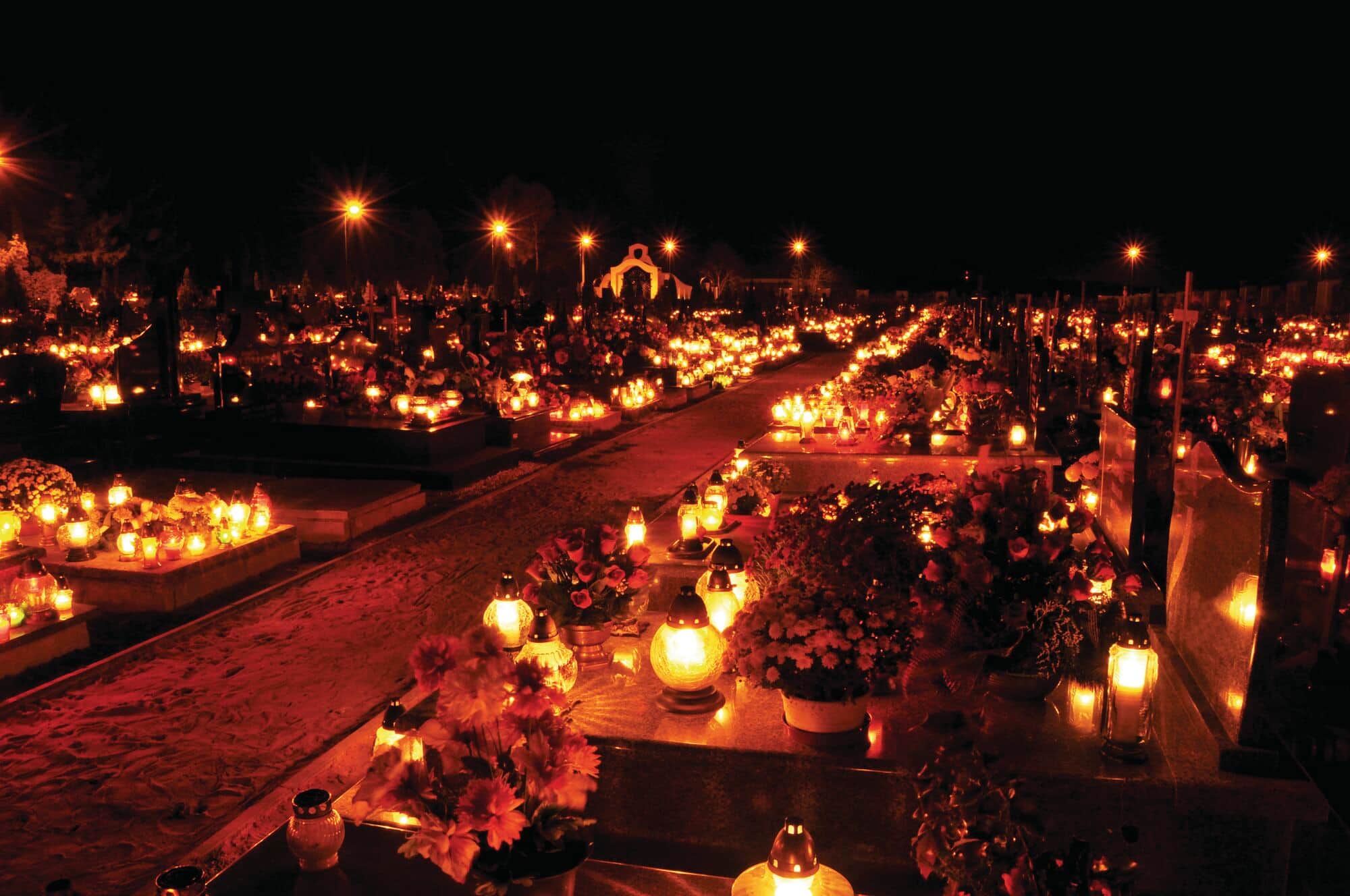 Lantern-lit cemetery with flower-covered graves at night.