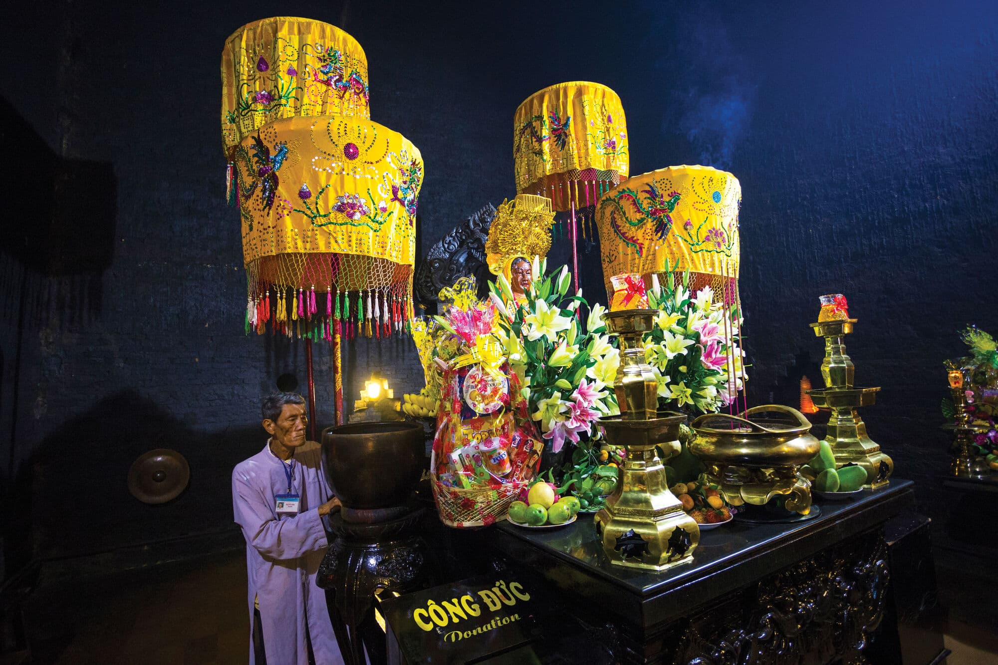 An altar adorned with flowers, fruit offerings and ornate yellow ceremonial canopies stands inside a dimly lit temple, with traditional decorative objects displayed around it.