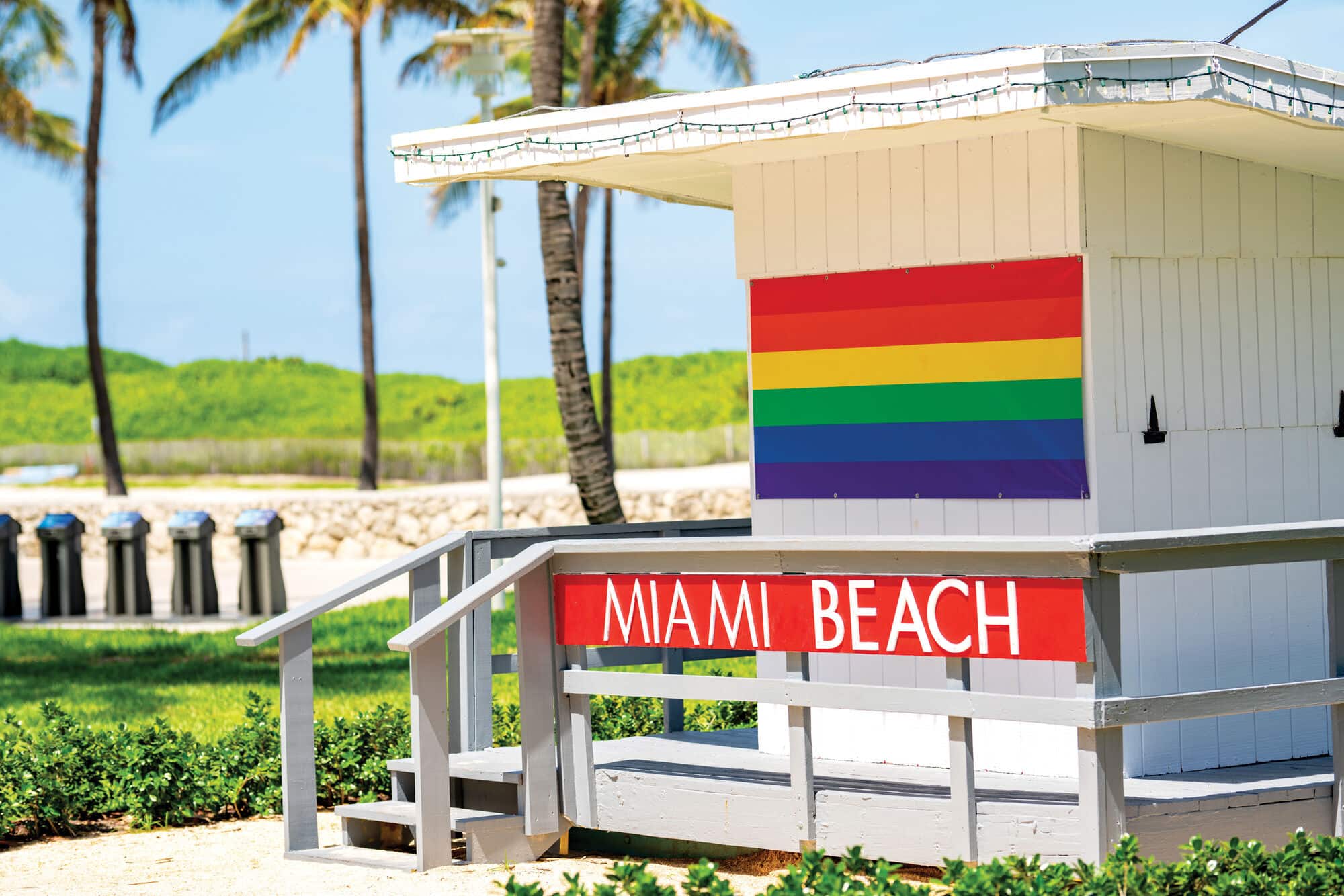 White beach hut displaying a rainbow pride flag and a red “Miami Beach” sign, with palm trees, sandy pathways and greenery in the background.