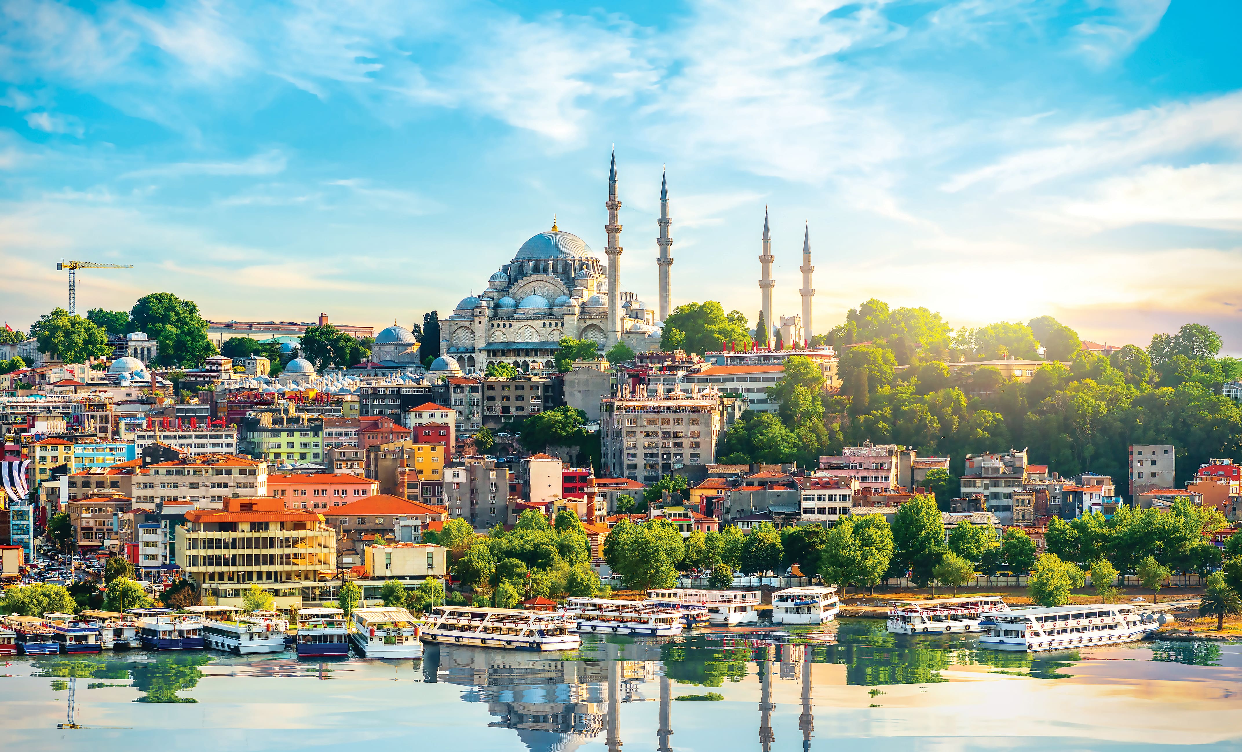 Panoramic view of Istanbul’s waterfront at sunrise, with the domes and minarets of a grand mosque overlooking a hillside of colourful buildings. Boats line the waterfront below, while the calm water reflects the city skyline beneath a bright blue sky.