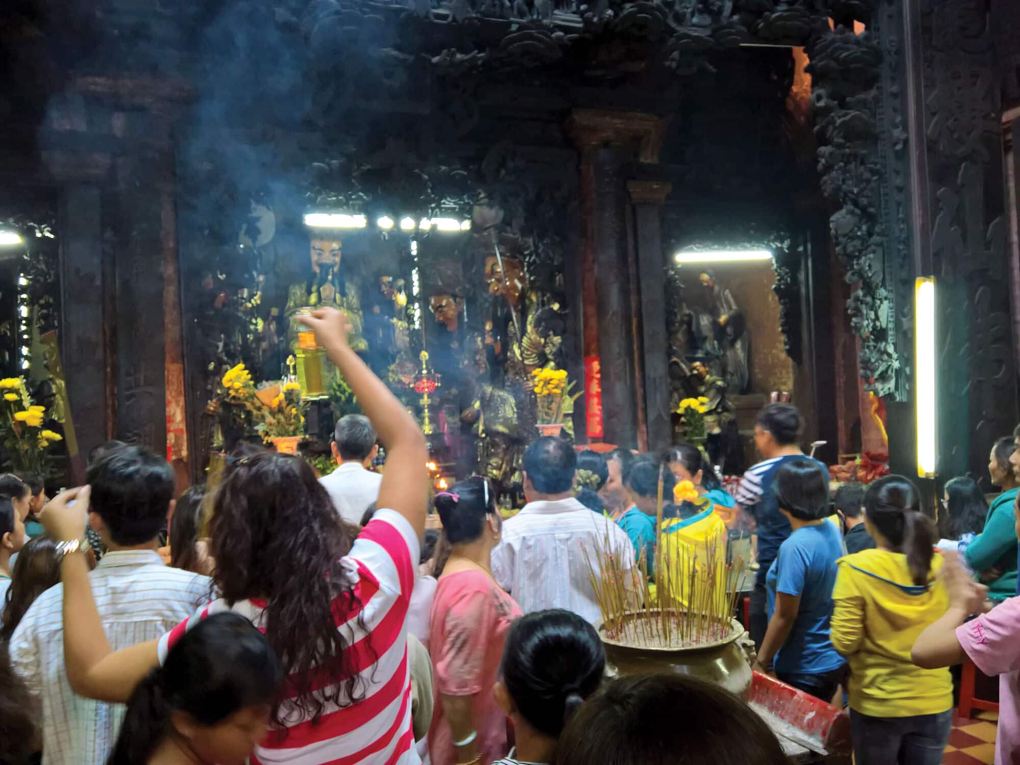 Visitors gather inside an ornate temple filled with incense smoke, carved pillars and glowing shrines, while offerings and incense sticks are placed in the foreground.