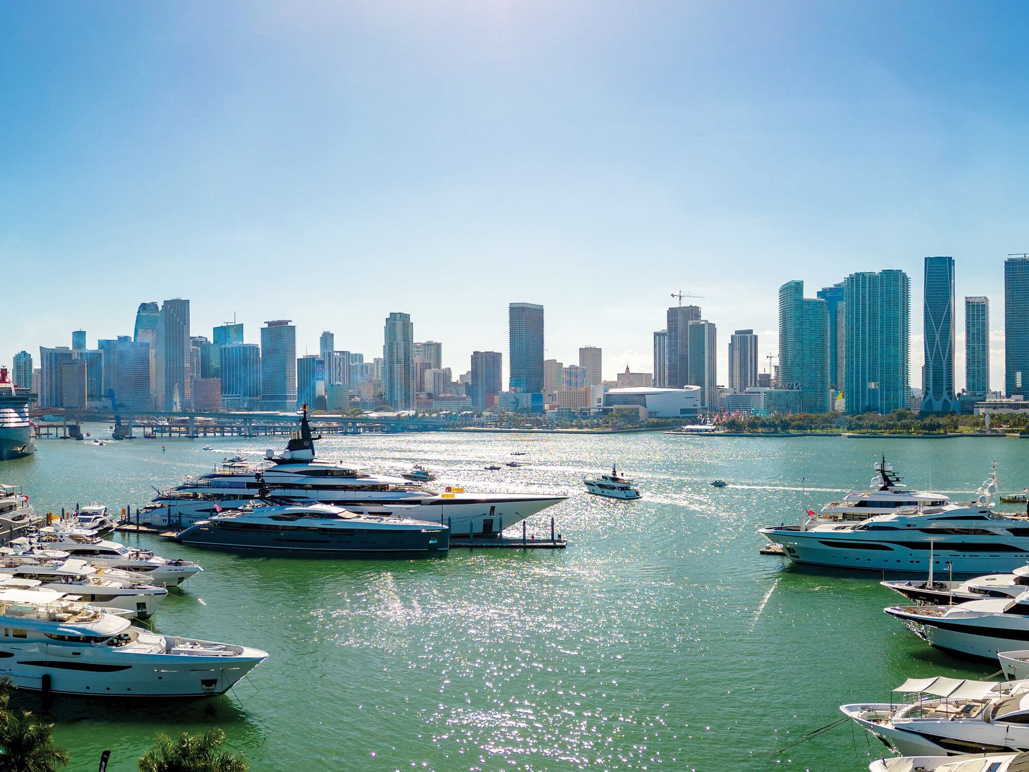 Luxury yachts moored in a marina overlooking a waterfront city skyline, with sunlight sparkling on the water and modern high-rise buildings in the background.