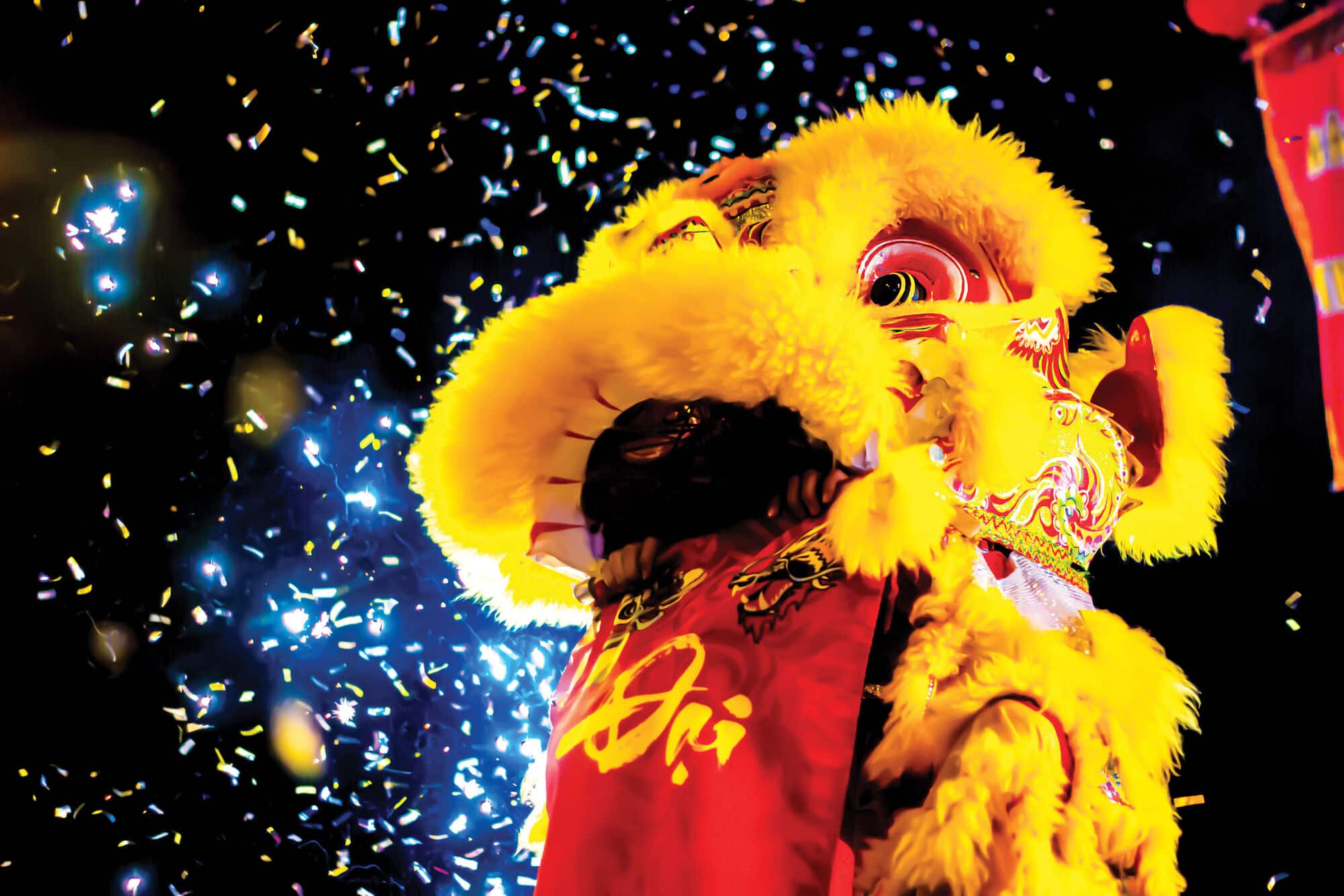 Bright yellow and red lion dance costume illuminated against a dark background, surrounded by sparkling confetti and lights during a festive cultural celebration.