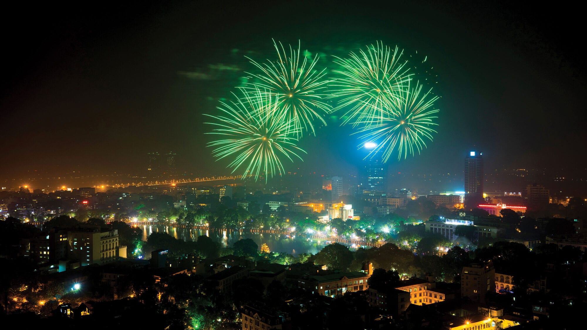 Green fireworks burst across the night sky above a brightly lit city, illuminating buildings and reflecting over a lake during a festive celebration.