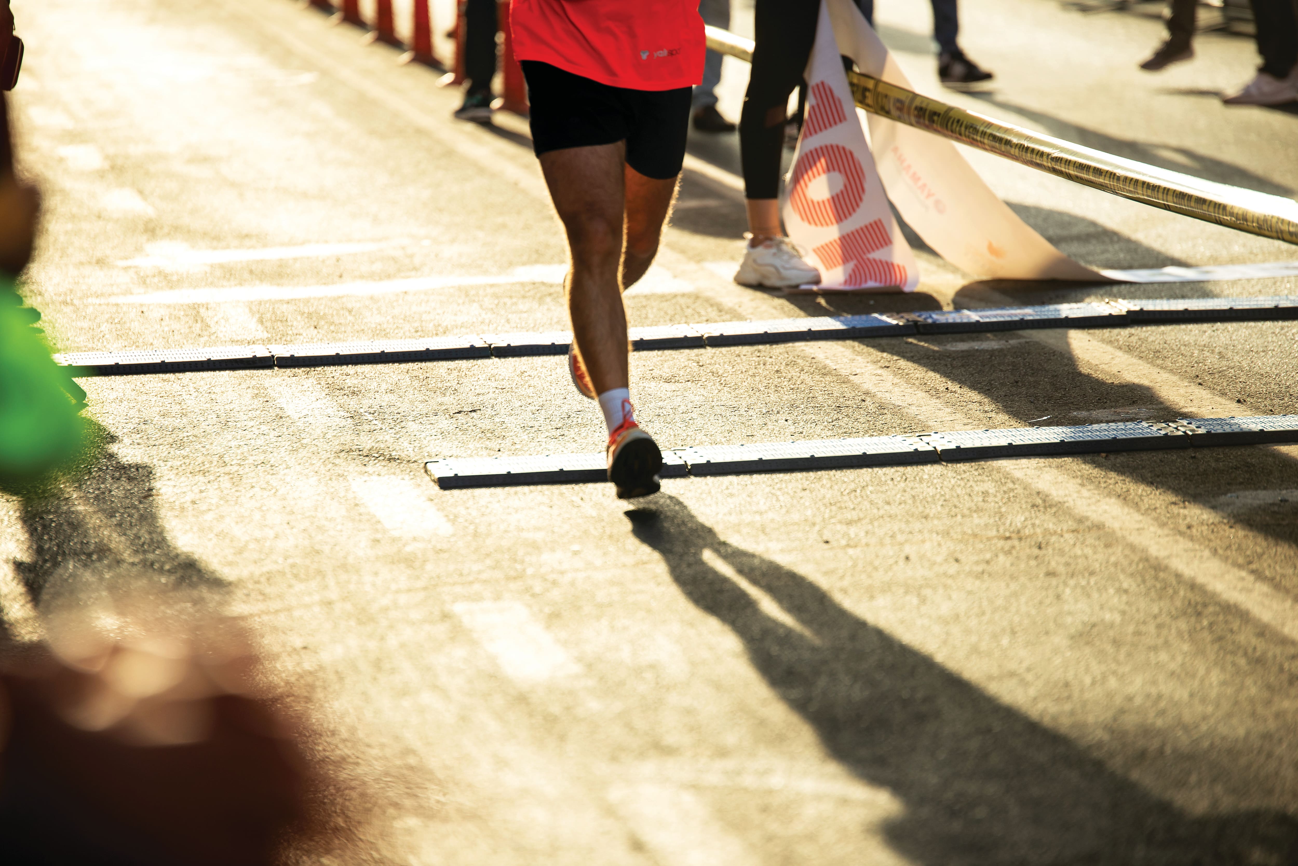 Close-up of a runner crossing a marathon finish line on a sunlit road. The athlete’s legs and running shoes are in focus, with timing mats stretched across the course and spectators standing behind barriers in the background.