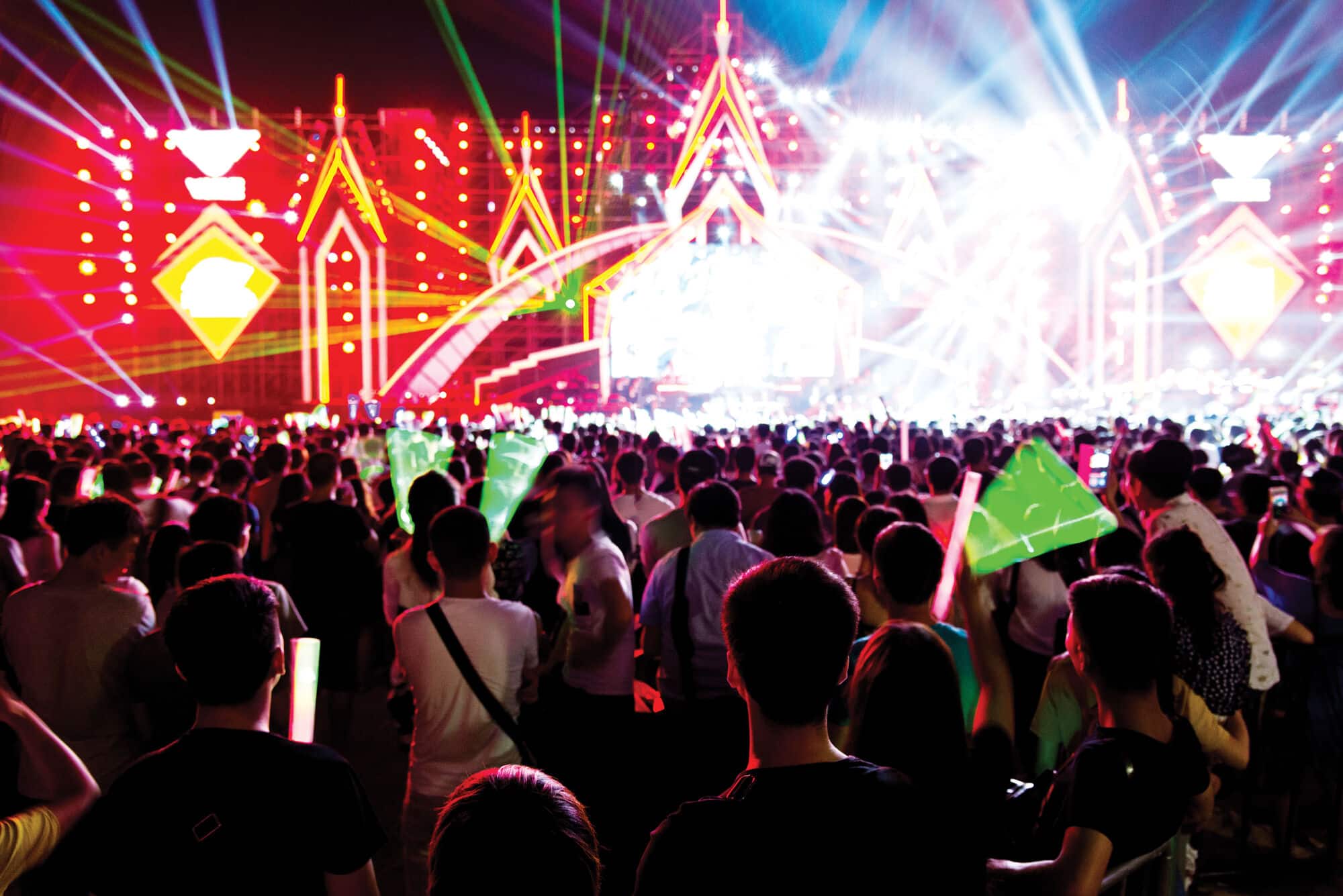 A large crowd gathers at a brightly lit outdoor festival or concert, with colourful stage lights, laser beams and illuminated decorations creating a vibrant nighttime scene.