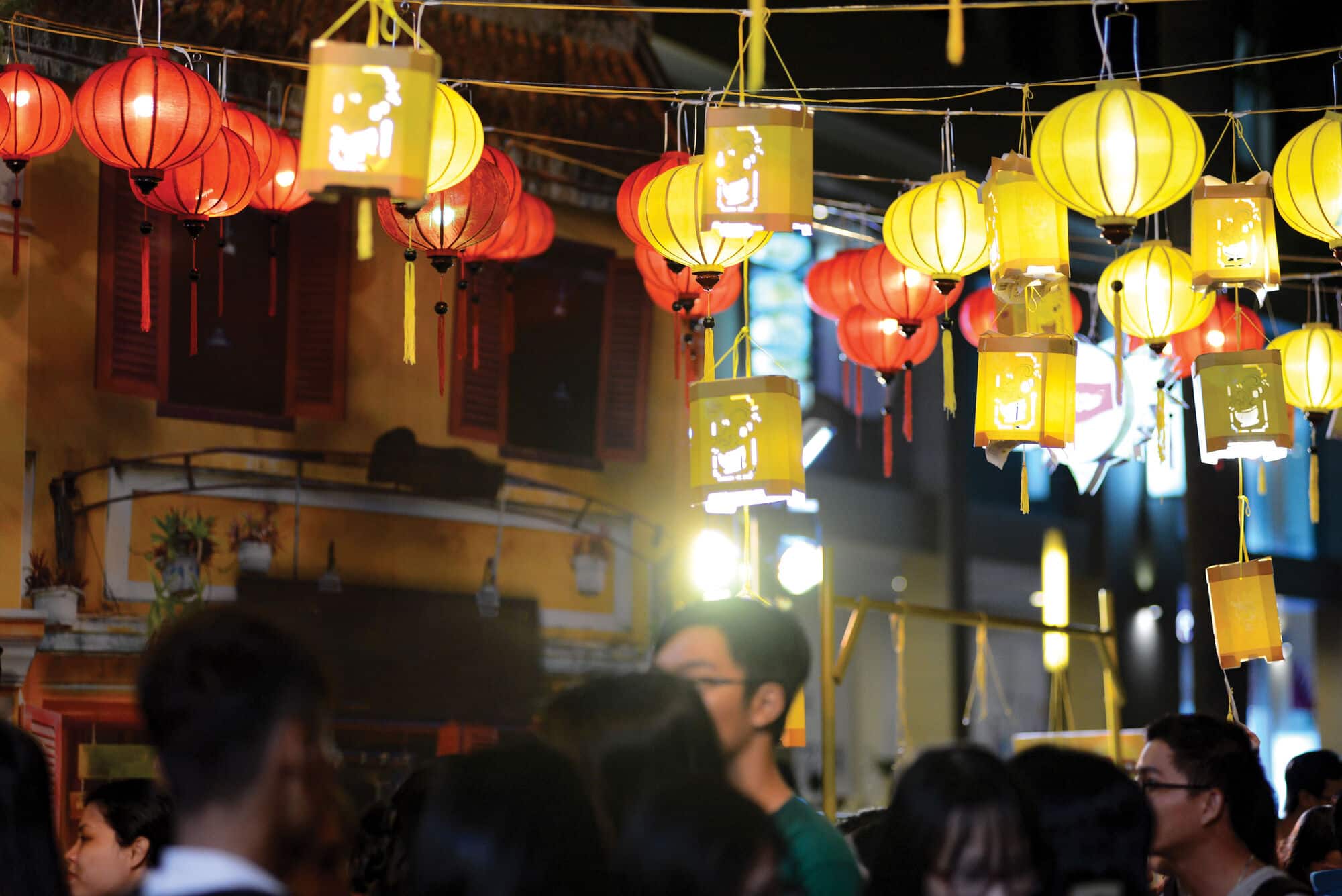 Red and yellow lanterns hang above a busy street at night, casting a warm glow over crowds below and creating a festive atmosphere among traditional buildings.