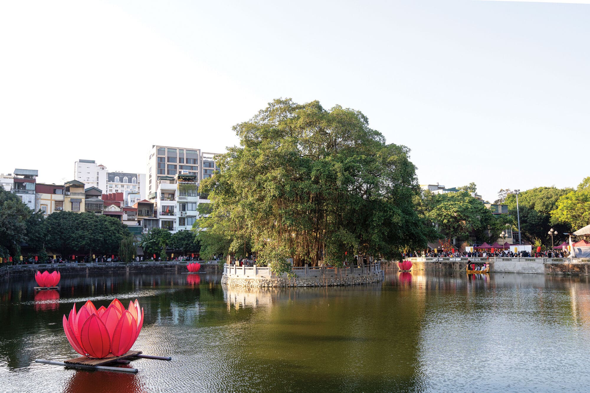 A calm lake surrounds a small tree-covered island, with large pink lotus sculptures floating on the water and city buildings visible in the background.