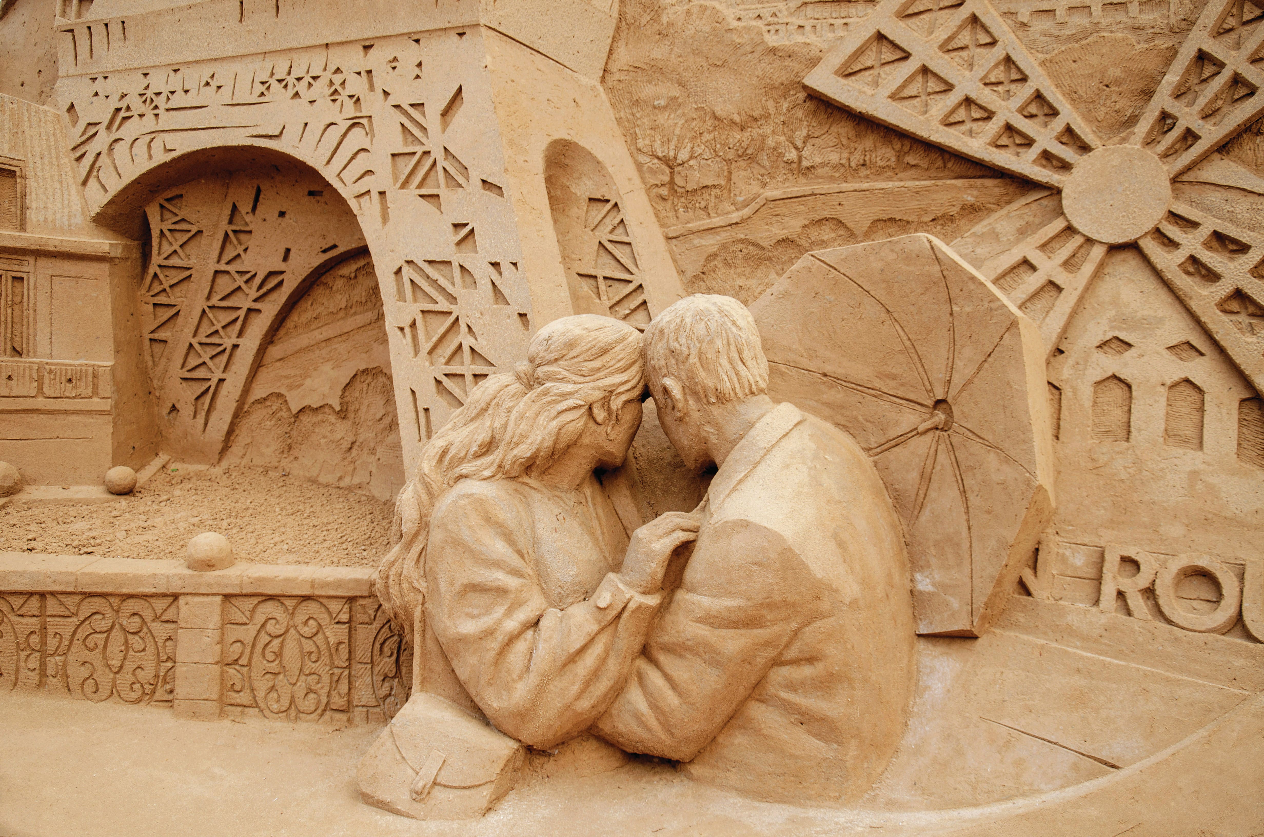 Intricate sand sculpture depicting a couple sitting together beneath an umbrella, surrounded by detailed architectural carvings and decorative patterns.