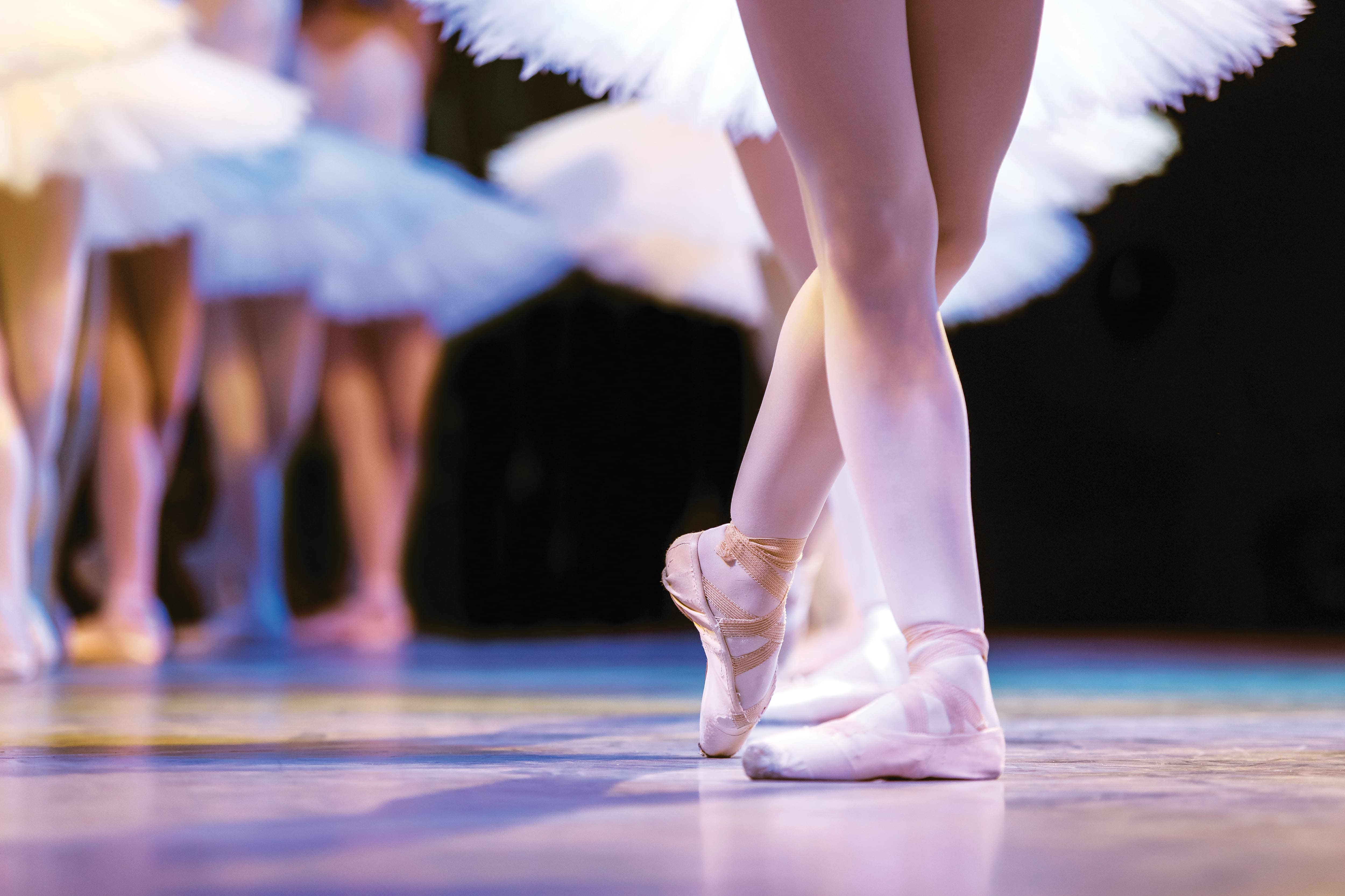 Close-up of ballet dancers performing on stage, with pointe shoes and crossed feet in sharp focus. White tutus blur softly in the background, while stage lighting casts a warm glow across the polished floor.