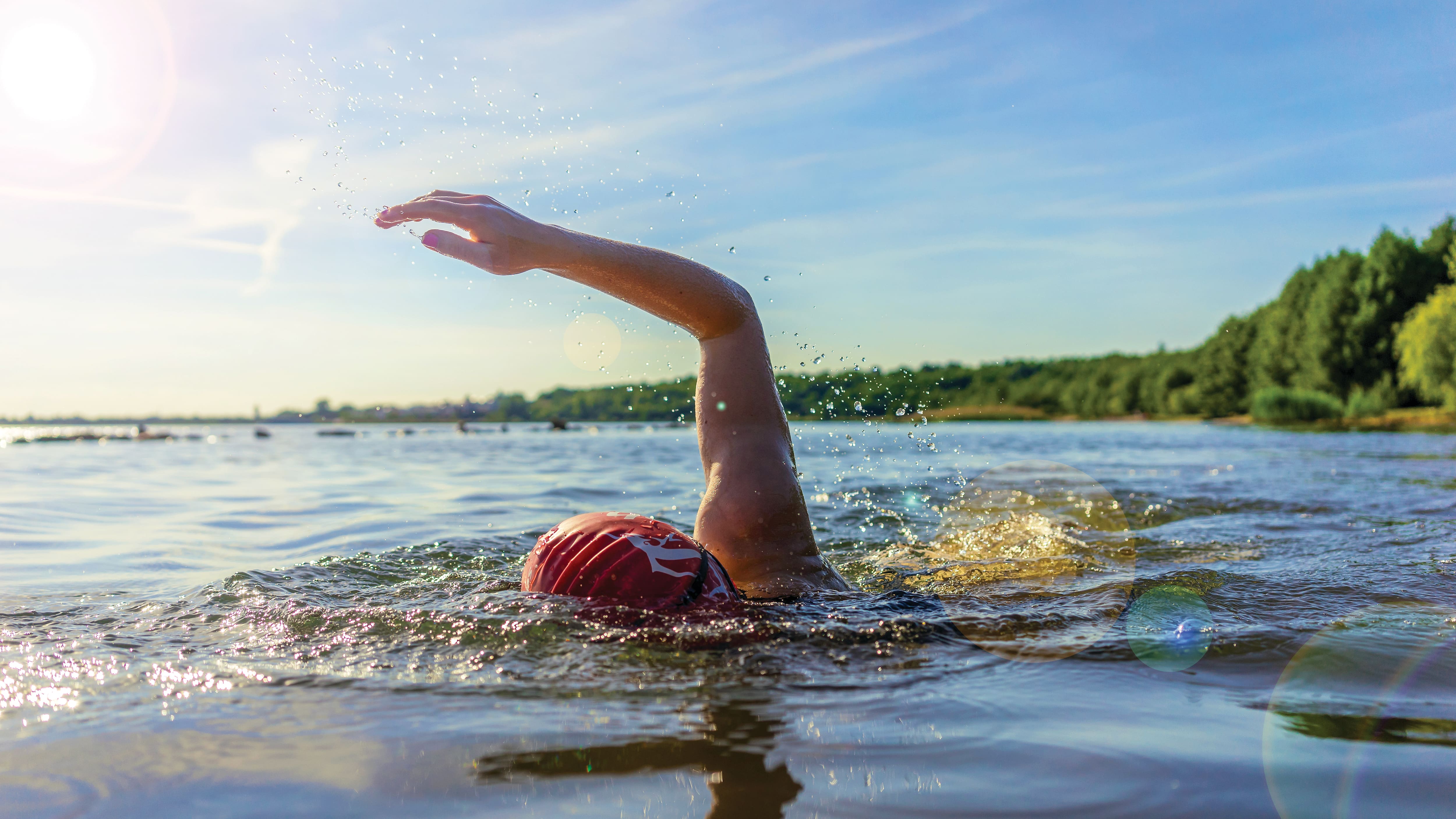 Open-water swimmer wearing a red swim cap performing a freestyle stroke across a sunlit lake. Water splashes around the raised arm, while a tree-lined shoreline and bright blue sky form the backdrop. 