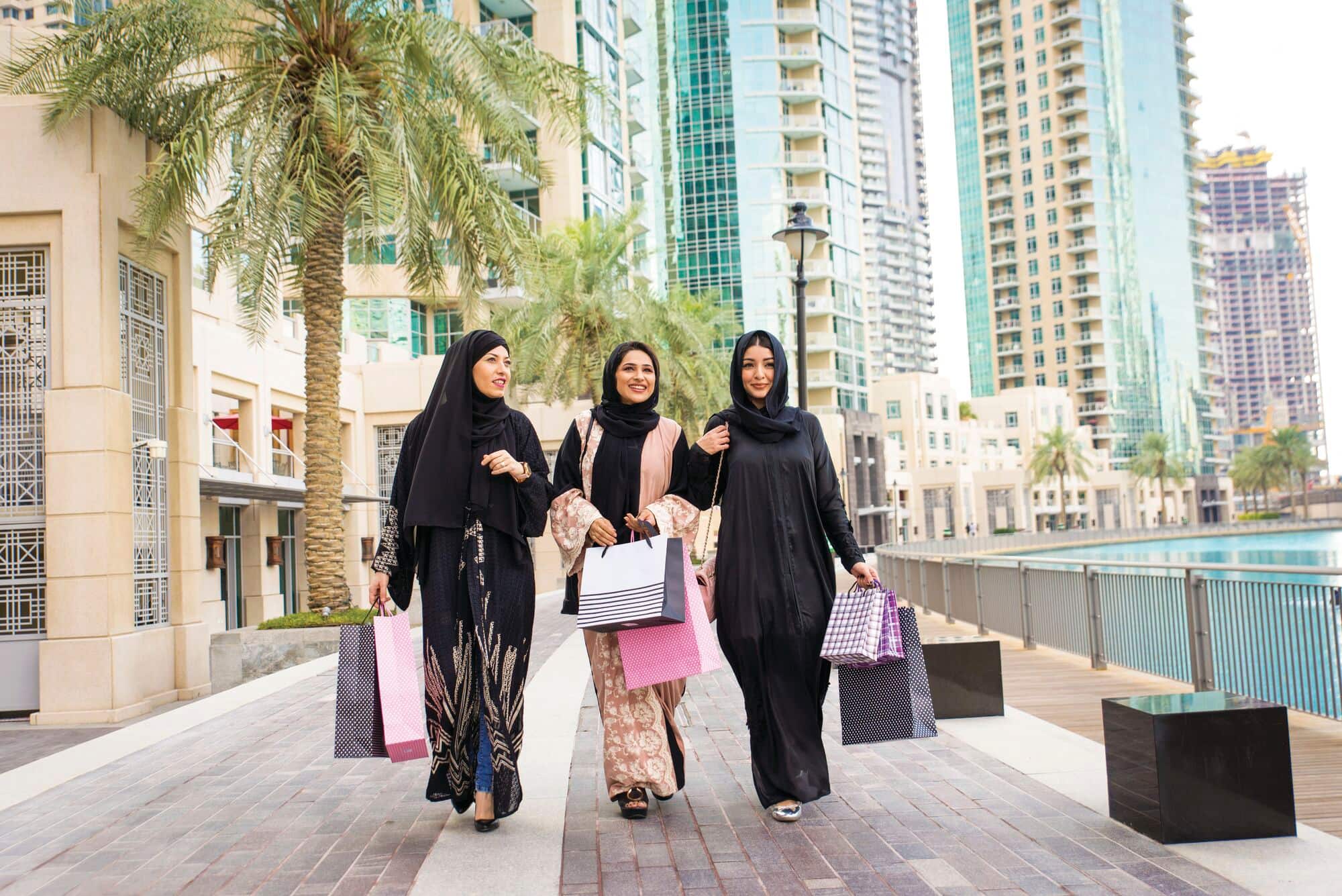 Three females walking outdoors, in Dubai, while wearing traditional abaya garments and carrying shopping bags