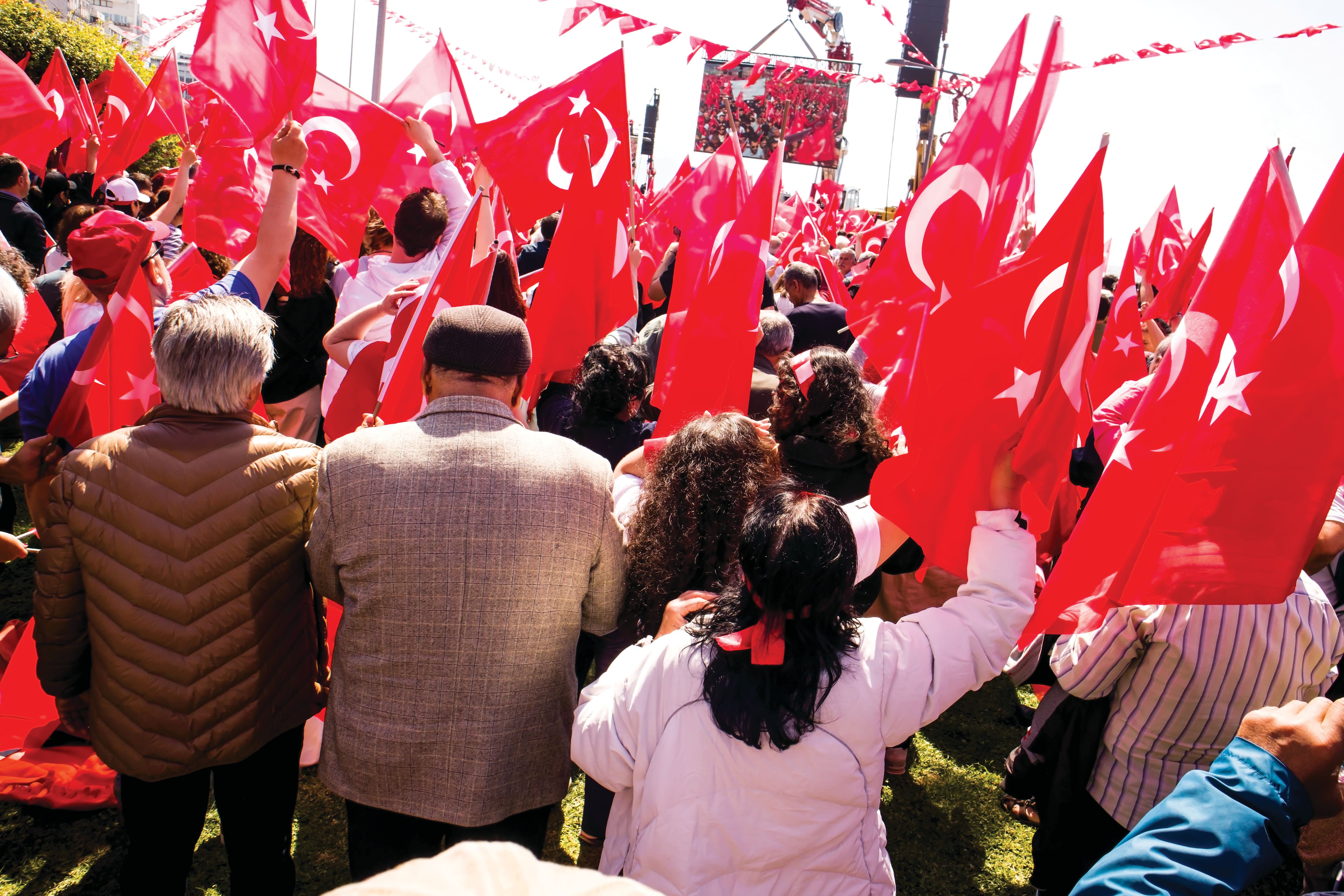 Large crowd gathered at an outdoor celebration, waving numerous red Turkish flags decorated with white stars and crescents. 