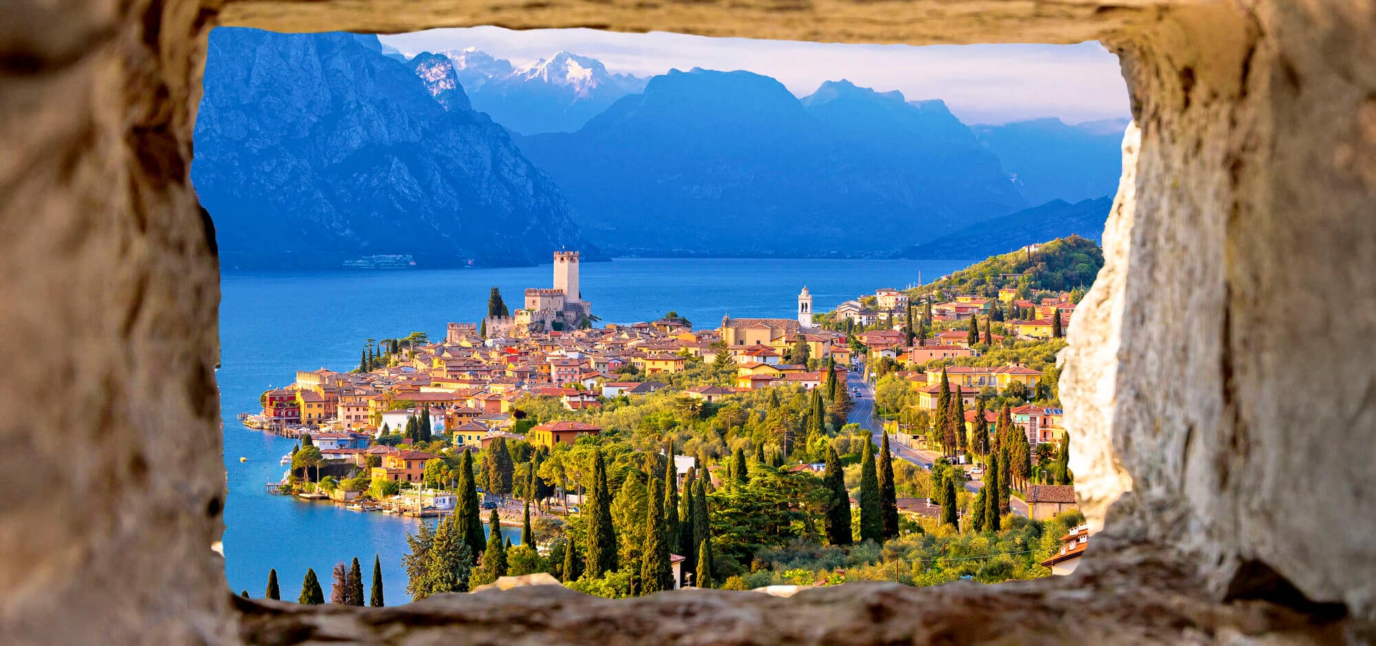  Colourful lakeside village with terracotta and pastel buildings, small boats moored in turquoise waters, backed by dramatic cliffs.